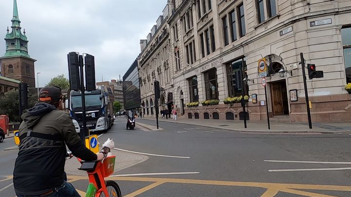 Sigrid, a white cat, rides away from camera in the basket of a Lime bike, being ridden by Travis, who wears a coat and a cap.