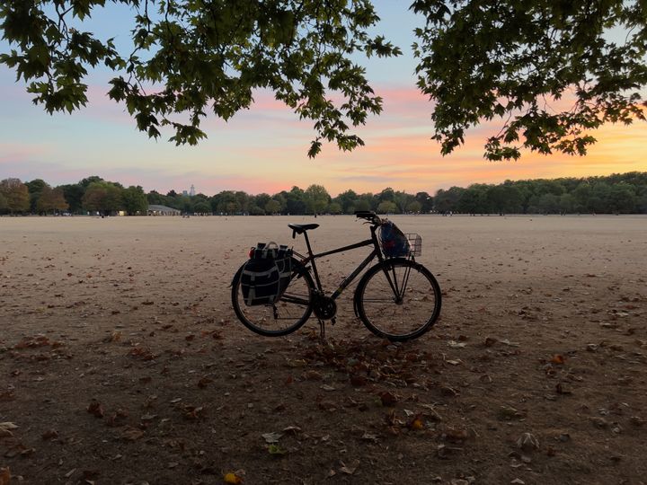 A bicycle on its kickstand in grass that is short and has dried out and turned brown, with a sunset in the distance.