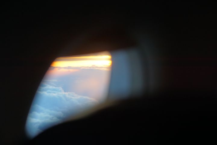 View of a pale yellow sunset over clouds lit in grey-blue through a plane window.