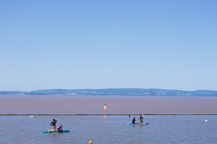 A person with orange shorts stands on a sea wall looking into a pool whose water is bluer than the brown Bristol Channel