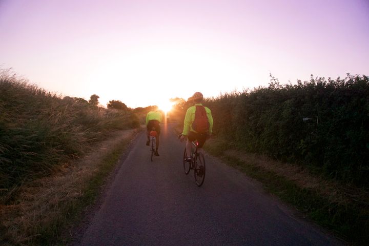 From the point of view of a cyclist: two other cyclists ride along a country lane into a purple-pink-gold sunrise.